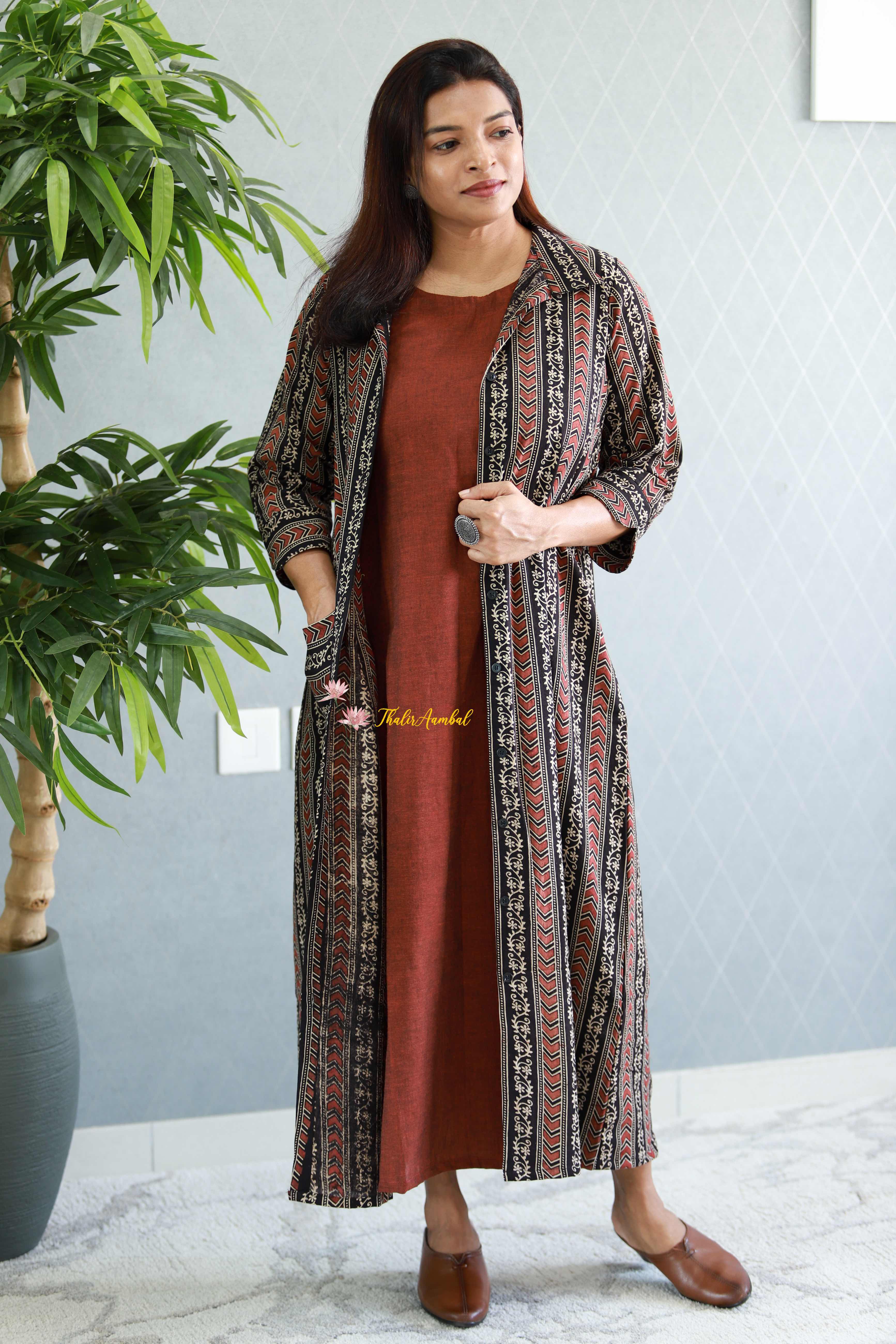 Woman wearing a long, patterned jacket over a rust-colored dress in a room with a plant and light gray wall. Thaliraambal