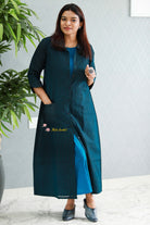 Woman wearing a long blue dress with a brand logo, standing indoors with a plant in the background. Thaliraambal