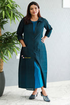 Woman wearing a teal dress with a gradient effect, standing indoors next to a plant. Thaliraabal
