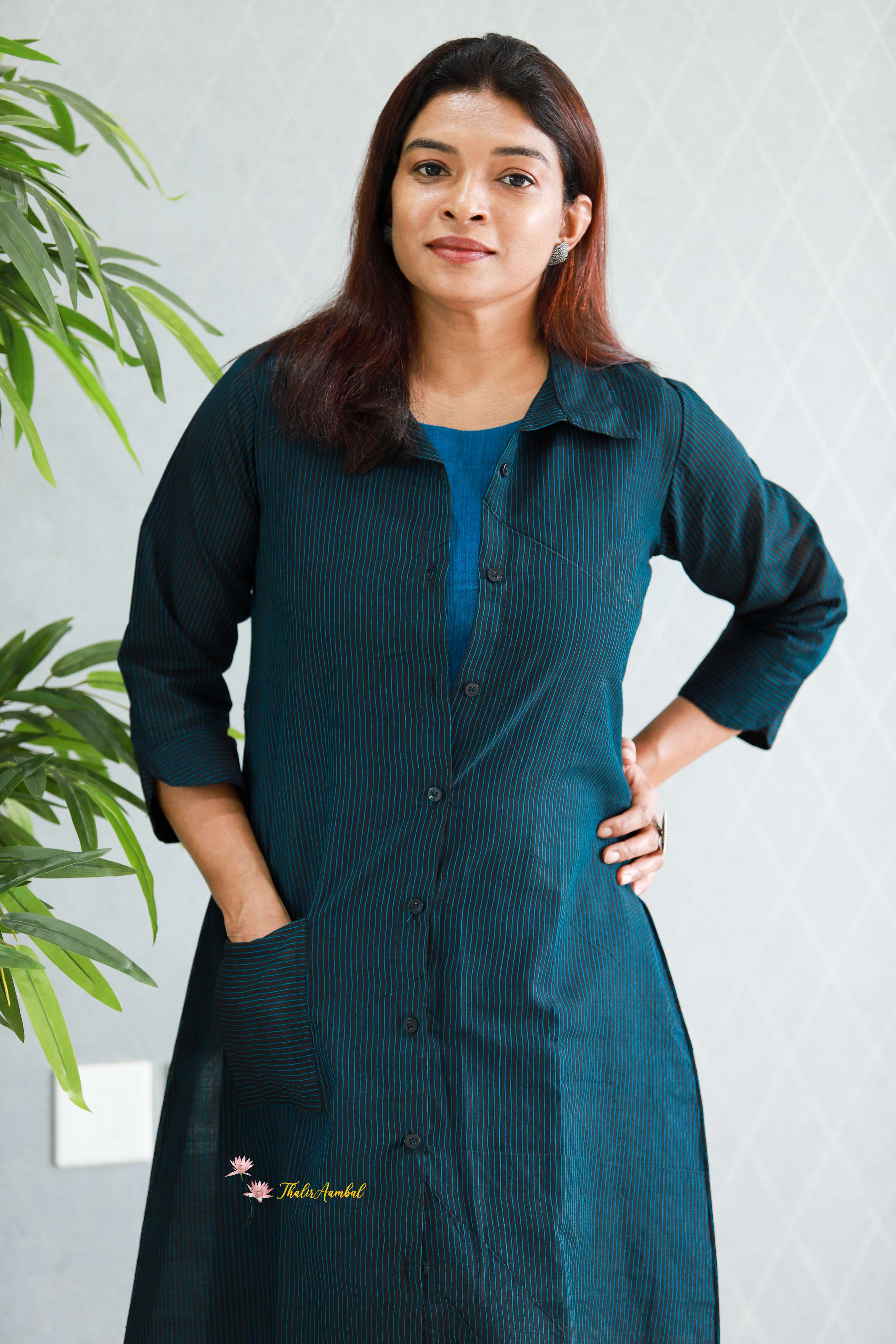 Woman wearing a teal dress with a white wall and plant in the background. Thaliraambal