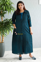 Woman wearing a teal black dress standing indoors with a plant in the background. Thaliraambal