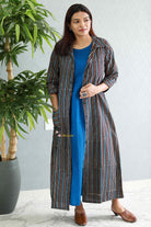 Woman wearing a long striped coat over a blue dress, standing indoors with a plant in the background.Thaliraambal