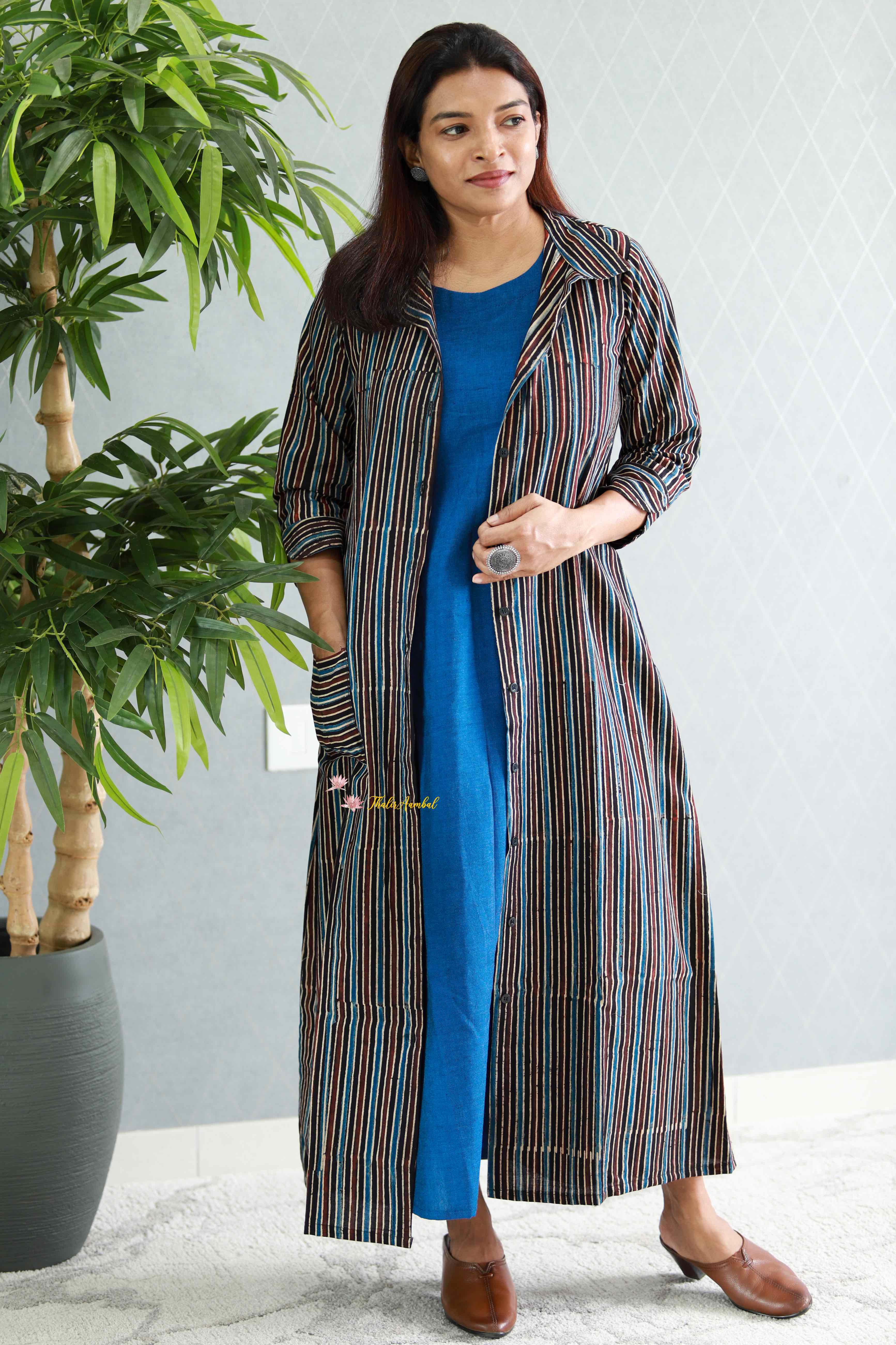 Woman wearing a long striped coat over a blue dress, standing indoors with a plant in the background.Thaliraambal