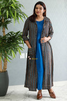 Woman wearing a blue dress with a long striped coat in a room with a plant.-Thaliraambal