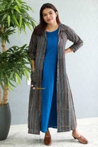 Woman wearing a long striped coat over a blue dress with a plant in the background.Thaliraambal