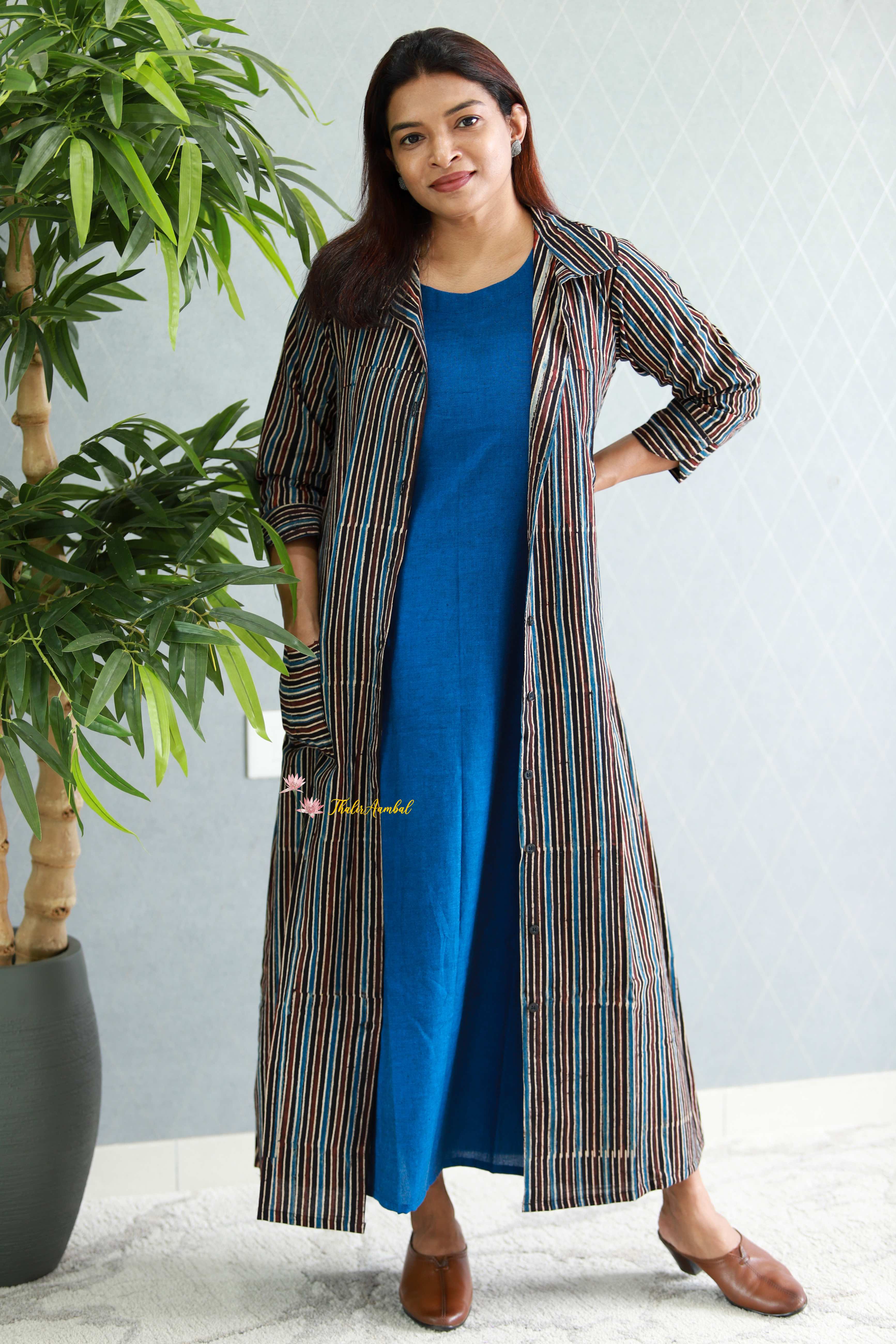 Woman wearing a long striped coat over a blue dress with a plant in the background.Thaliraambal