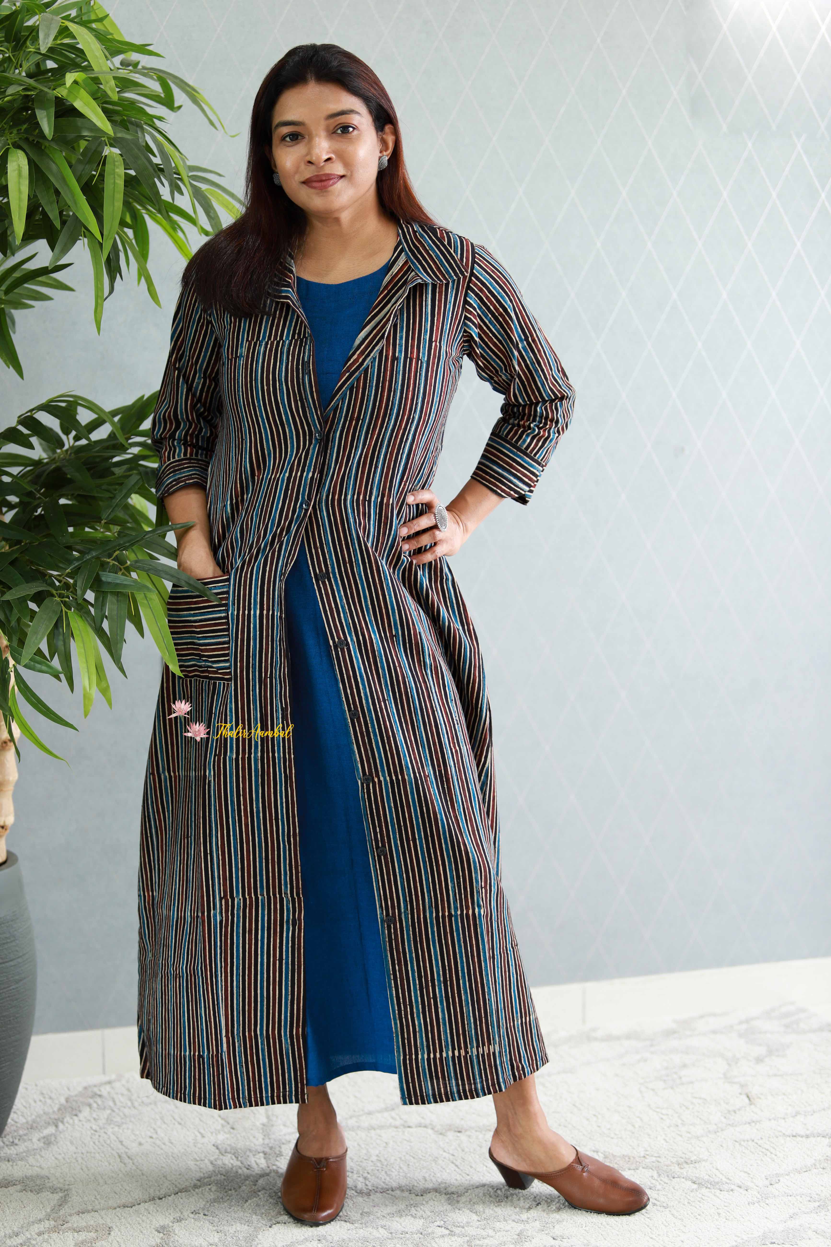 Woman wearing a long, patterned coat over a blue dress, standing indoors with a plant in the background.Thaliraambal