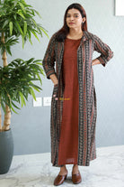 Woman wearing a long patterned coat over a brown dress indoors with a plant in the background. Thaliraambal