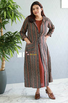 Woman wearing a patterned long coat over a red dress, standing indoors with a plant in the background. Thaliraambal