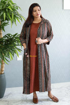Woman wearing a long, patterned jacket over a rust-colored dress in a room with a plant and light gray wall. Thaliraambal