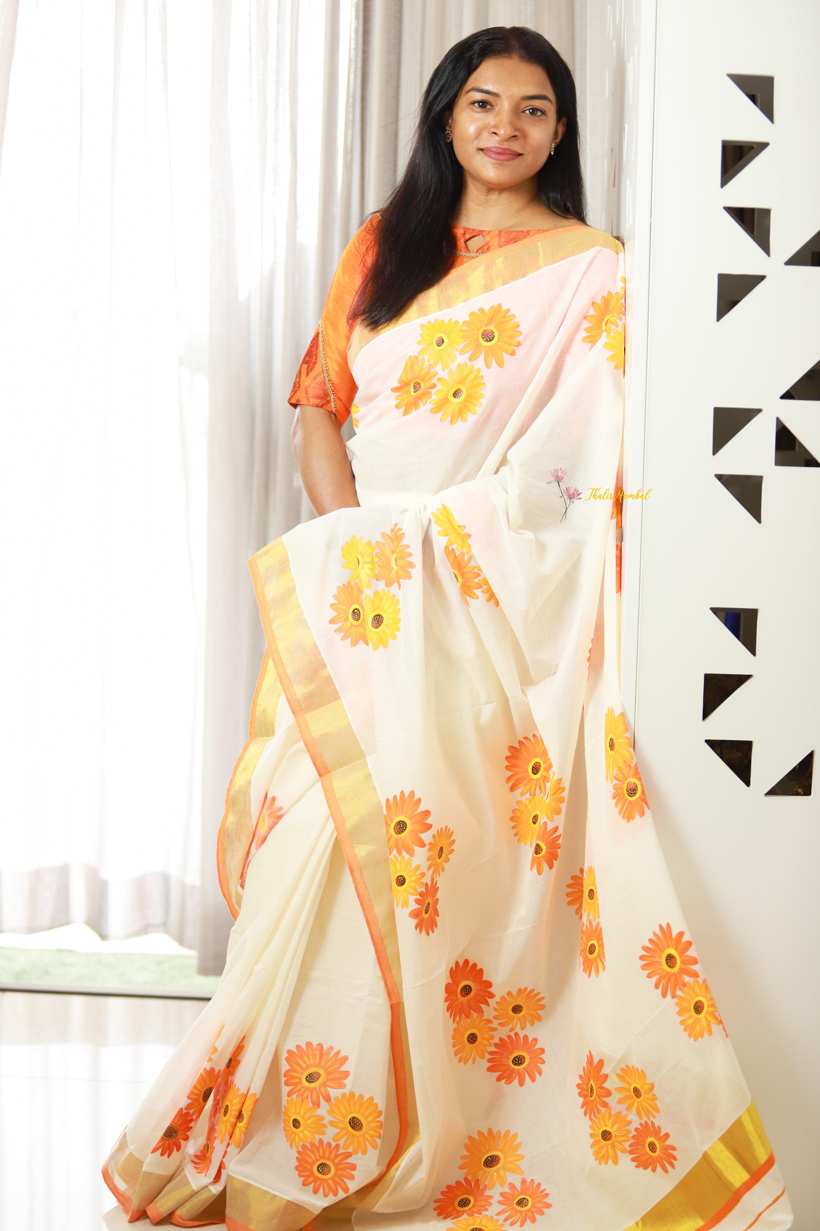 Woman wearing a white saree with orange and yellow floral patterns, standing indoors.Thaliraambal